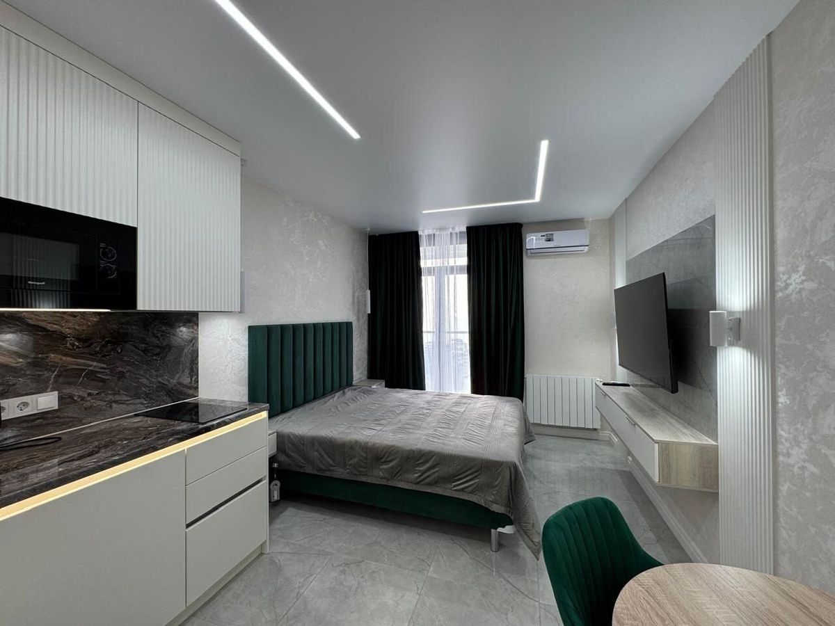 Kitchen unit with a dark splashback and a bed with a green headboard sharing one room